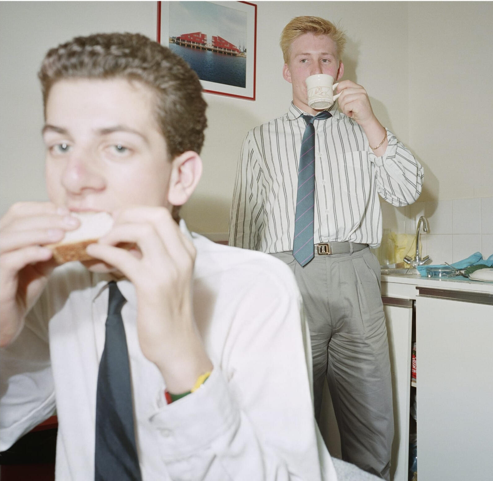 Two young men in shirt and ties enjoy tea and a sandwich in a corporate breakroom in the 1980s. Photo by Anna Fox (1988)